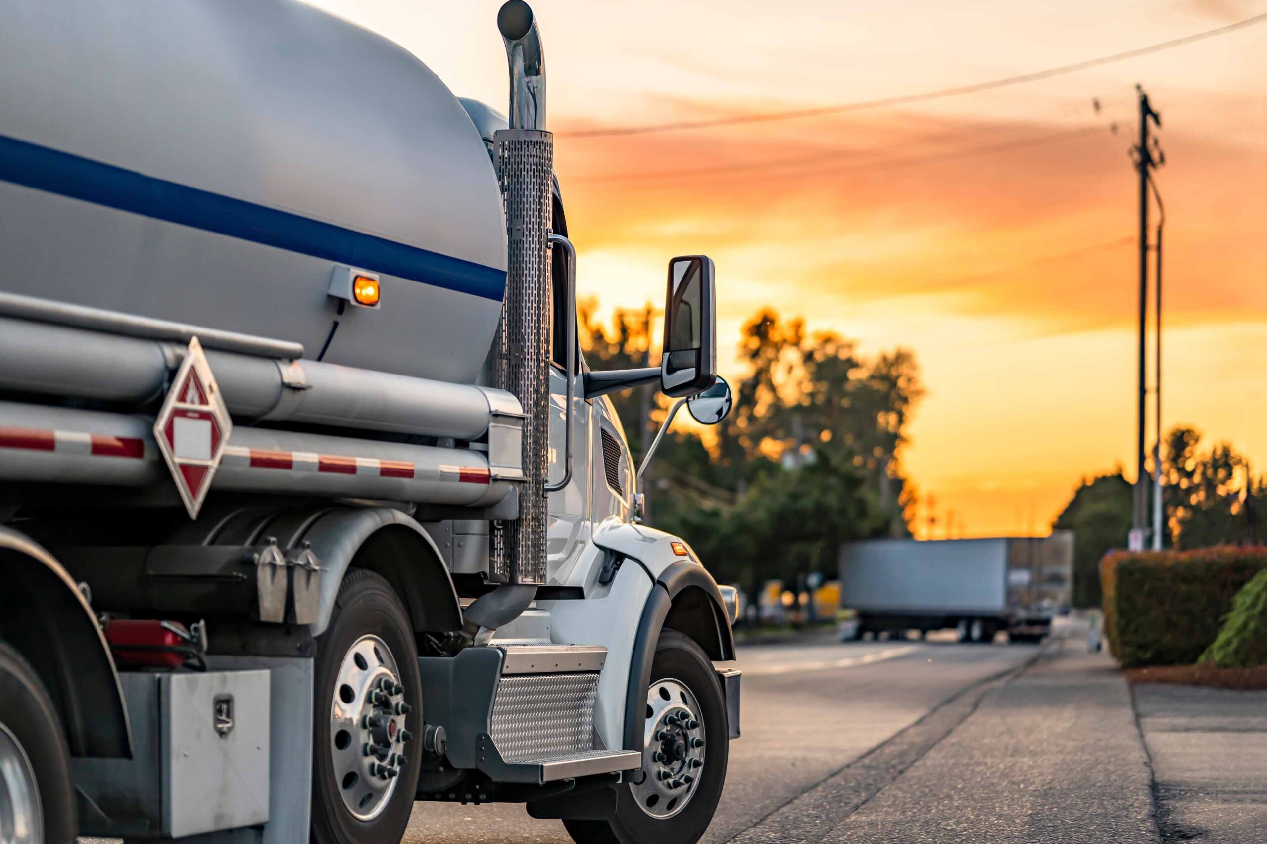 A large tanker truck is driving on a road at sunset. The truck has a cylindrical tank with a blue stripe and hazard placards on its side. In the background, there is another vehicle, possibly a trailer, and trees lining the road. The sky is filled with orange and yellow hues from the setting sun.
