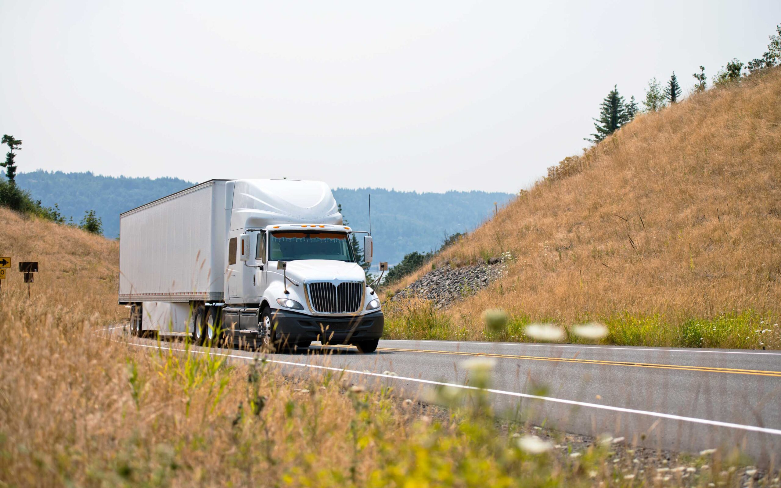 A white semi-truck with a trailer drives along a two-lane rural road bordered by dry grass and rolling hills, under a clear sky with forested areas in the background.
