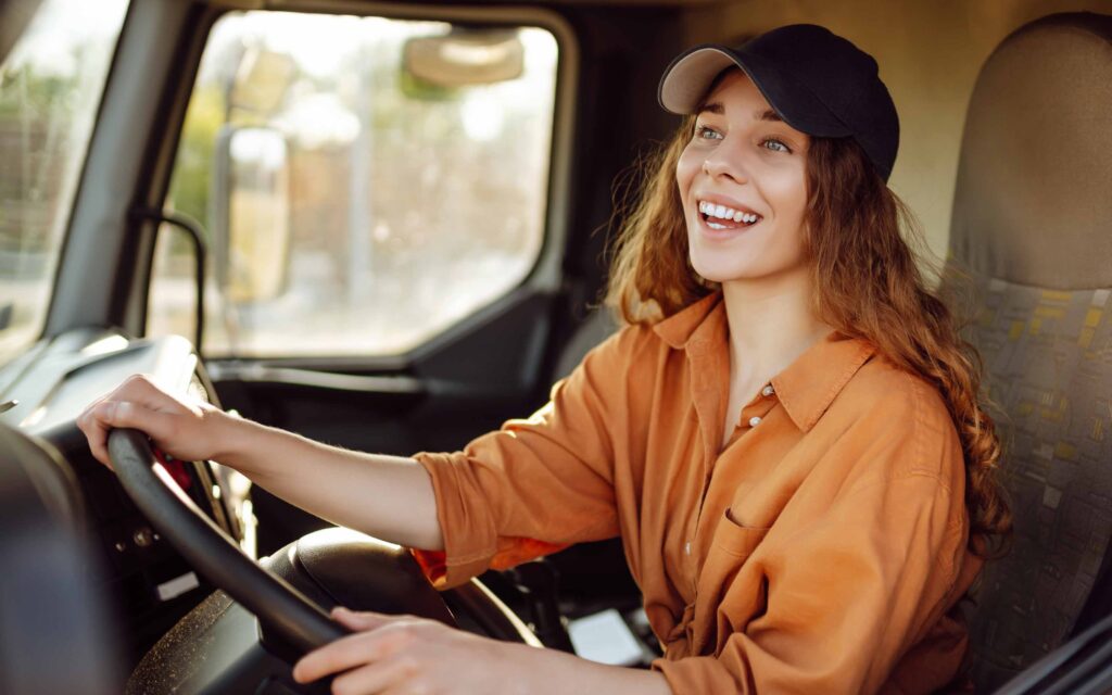 A smiling female truck driving student wearing a cap holds the steering wheel, happy during her training in the truck's cab.
